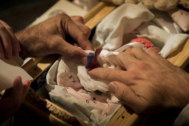 Mains d'homme et de femme tenant entre leurs doigts le minuscule pied d'un mort-né, dont la plante a été recouverte d'encre pour en prendre l'empreinte sur une feuille de papier.