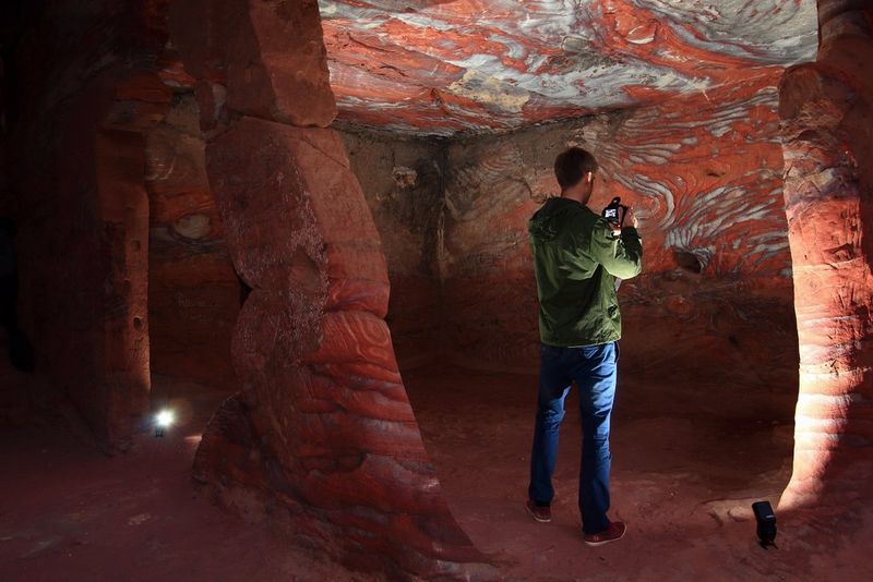 A photographer stands in a cave taking a picture of a rock wall with swirling patterns in it, illuminated by two off-camera flash units on the floor.