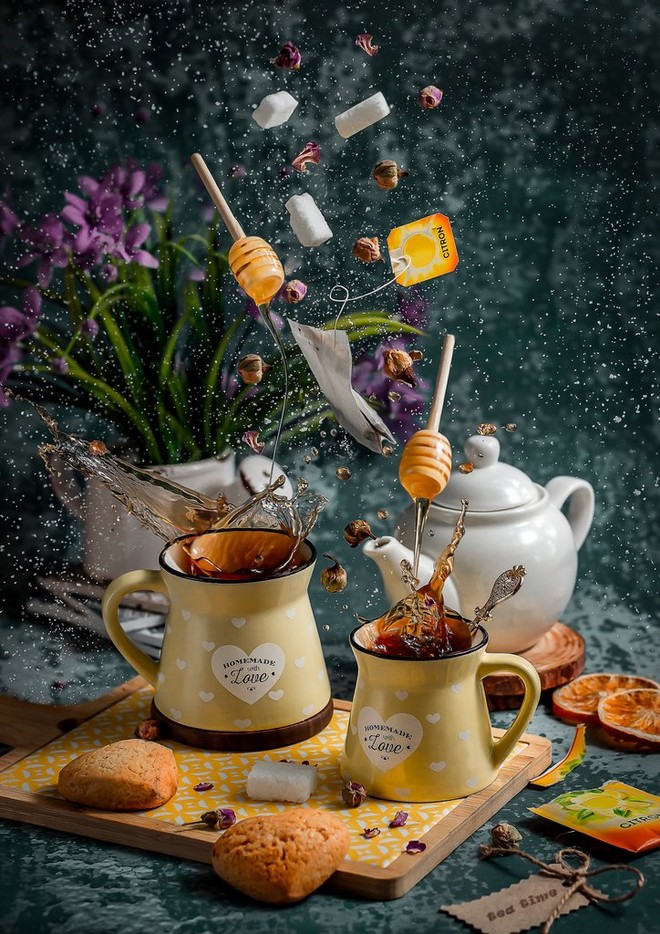 Zollette di zucchero, mestoli di miele e bustine di tè sospese sopra due tazze di tè; schizzi di liquido vengono congelati in movimento sopra i bordi delle tazze.