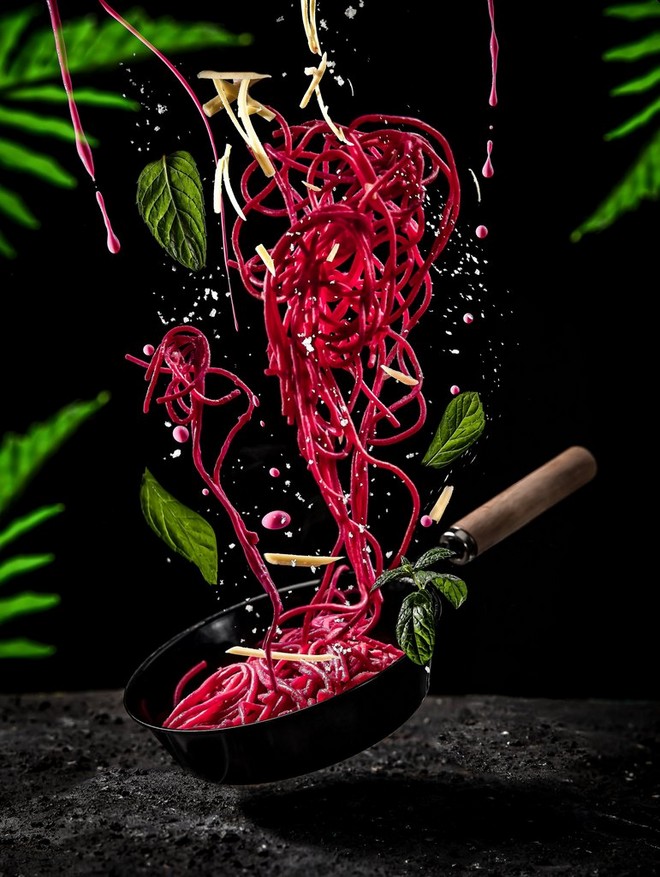 Spaghetti rosa che cadono in una padella sospesa sopra un bancone, davanti a uno sfondo nero e incorniciati da foglie verde brillante.