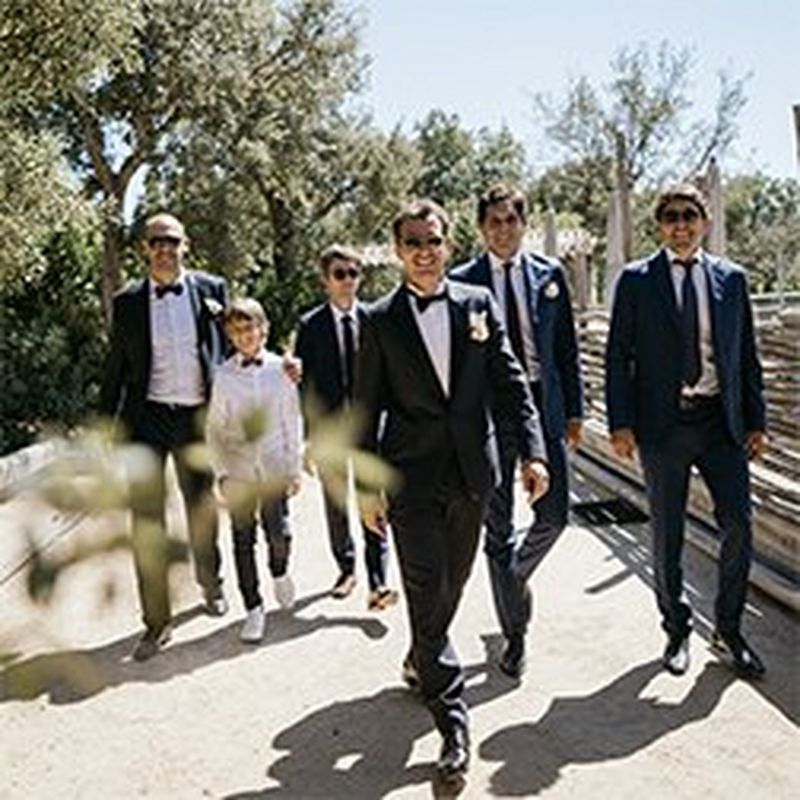 A groom and five groomsmen of various ages, most wearing sunglasses, walk towards the camera smiling broadly.