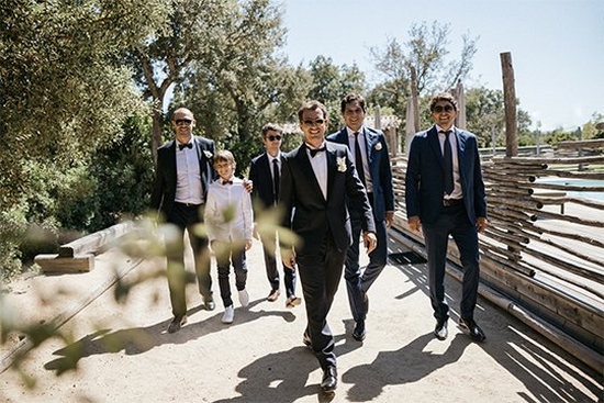A groom and five groomsmen of various ages, most wearing sunglasses, walk towards the camera smiling broadly.