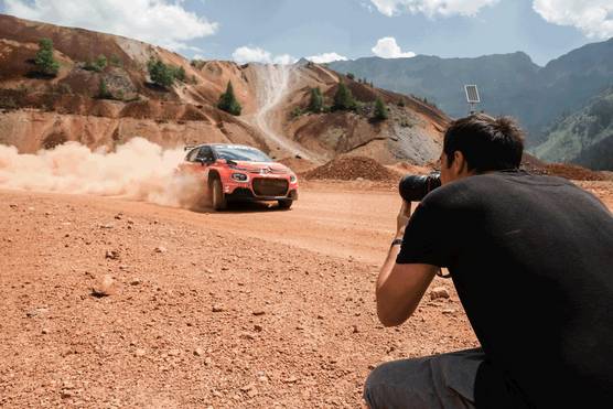A photographer crouches to take a shot as a red sports car speeds down a desert road, dust billowing behind it.