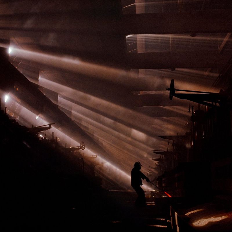 A person holds a hot steel rod, the end of which is glowing red. Only the silhouette of the person is visible as light creeps in through the many slits on the left side of the room. This photograph was captured by James Nachtwey in Czechoslovakia in 1990.