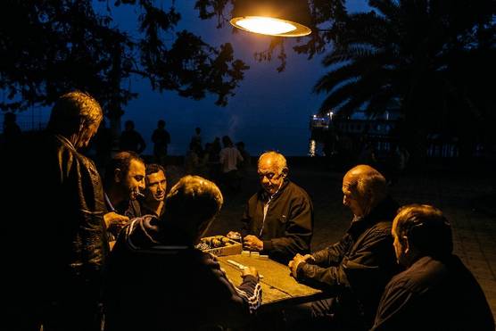A group of Abkhaz men sit around a table near the sea playing dominoes under a lamp.