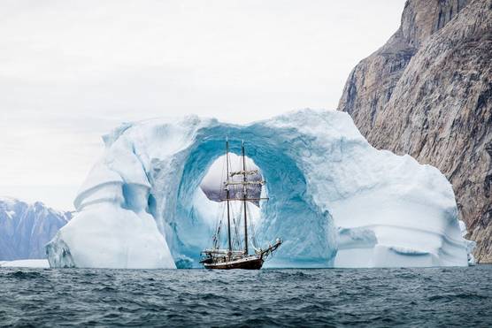 A large sail-powered ship passing through an archway in an iceberg on a rocky coastline. 