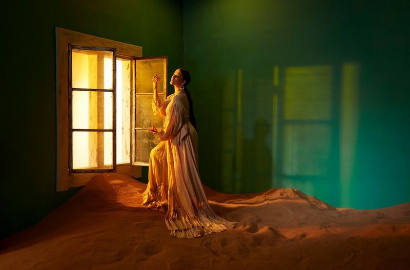 A woman in a white dress stands next to an open window which lets light into a room with a green wall and sand on the floor. 