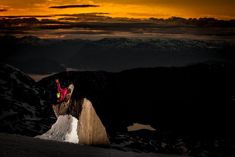 Uno snowboarder esegue un salto su una sporgenza innevata, con un tramonto dorato sullo sfondo.
