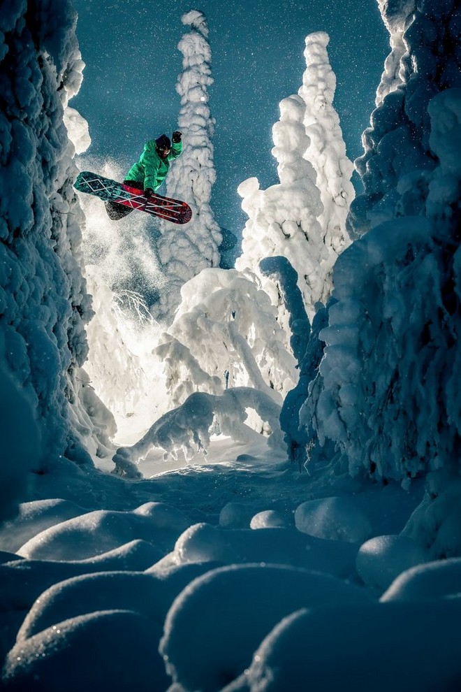 Сноубордист в яркой одежде во время прыжка, окруженный сказочными заснеженными деревьями.