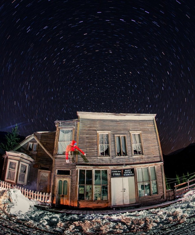 Деревянное сооружение «под старину» на фоне ночного неба со скругленными траекториями движения звезд; на переднем плане прозрачный сноубордист в красной кофте прыгает с фасада здания.