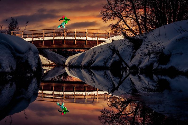 Сноубордист скользит по деревянным перилам моста между двух заснеженных берегов — мост идеально отражается от поверхности воды.