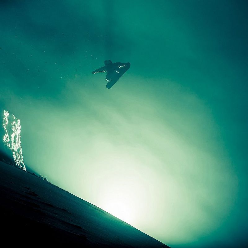 Ein Snowboarder bei Nebel, hoch in der Luft, an einer atmosphärisch von hinten beleuchteten Bergkuppe.