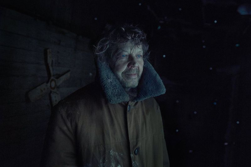 A man with bright blue eyes and curly hair, wearing a thick brown coat, standing indoors in the dark, lit only by the light from the window he is looking out of.