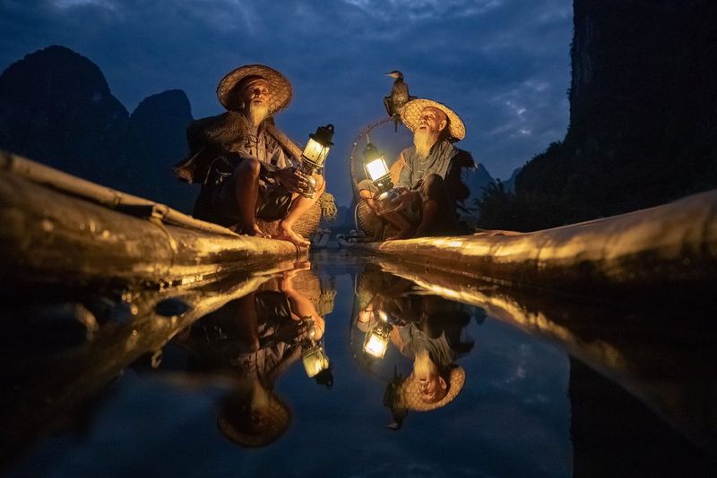 رجلان صينيان يعملان في صيد السمك بواسطة طائر الغاق يجلسان في قاربين طويلين ومعهما مصباحان.