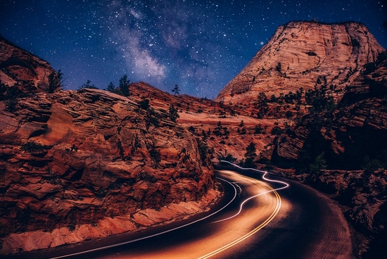 Rocky outcrops in Zion National park, Utah, USA, pictured at night. Blurred lights run along the road and the Milky Way can be seen in the sky above.