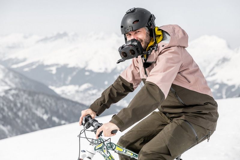 A man rides a bicycle in a snowy scene with steep, snow-covered mountains behind, wearing a helmet with a camera rigged to the front.