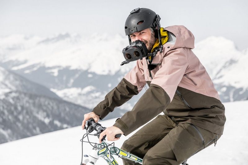 Un uomo va in bicicletta tra la neve, con montagne ripide e innevate sullo sfondo, indossando un casco con una fotocamera montata sulla parte anteriore.