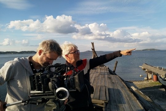Two men stand on a docked wooden boat with a large body of water behind them. One is adjusting a Canon EOS C500 Mark II camera, the other is pointing into the distance.