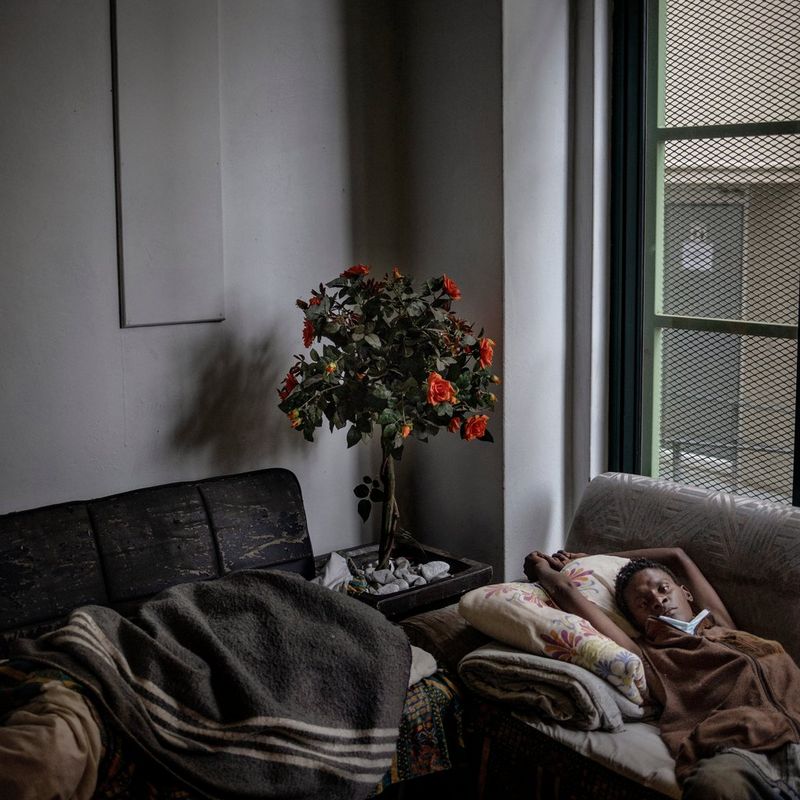  A woman lies across a sofa with her hands draped above her head, a face mask pulled down to her chin. In the corner of the grey room is a small planted tree with bright orange flowers. 