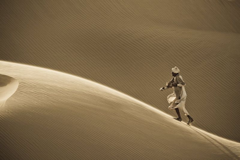 A man runs up a sand dune in India's Thar Desert, a huge expanse of rippling sand behind him, in a minimalist portrait by Joel Santos.