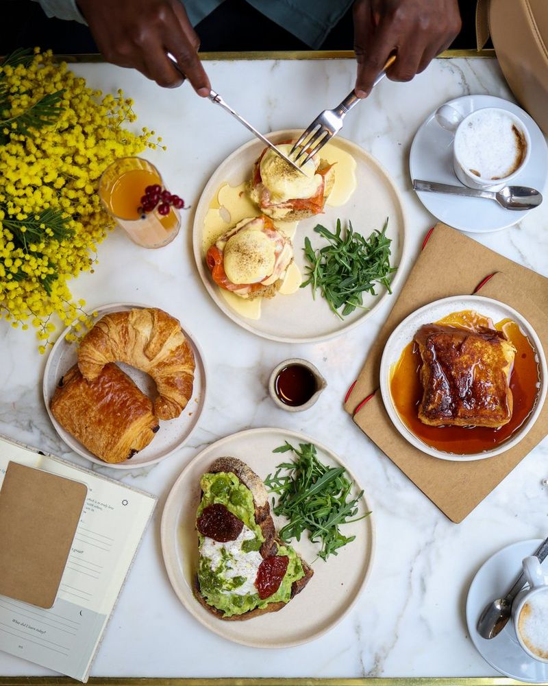 A top-down view taken by Laura Hannoun of a café table, showing four plates of fresh food, drinks, flowers and notebooks. A pair of hands is holding a knife and fork and starting to cut into one of the meals.