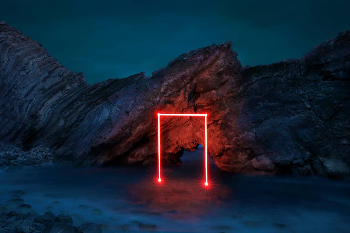 A multiple exposure image of a doorframe outlined in red light. The shape appears to emerge from the sea and is set against a rocky backdrop.