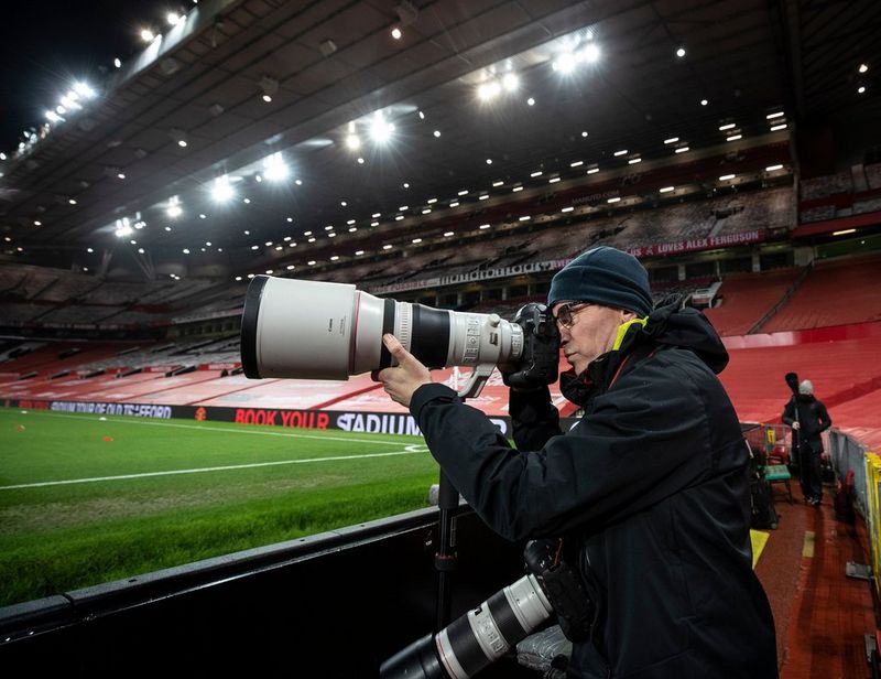 Эдди Кио на стадионе; он держит камеру с большим объективом, а другая камера с объективом висит у него на шее.