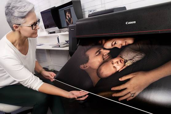 In an editing studio, a large print showing a baby, mother and father is coming out of a large format printer, while a smiling woman carefully holds up the edges of the print.