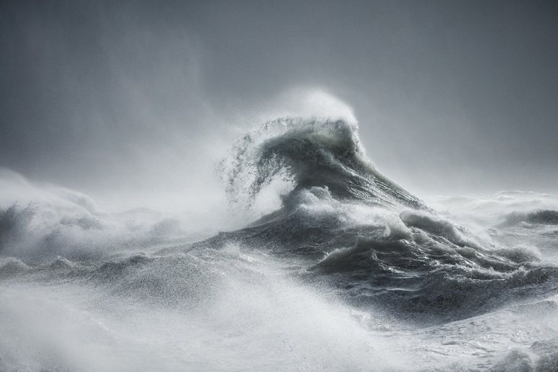 A black-and-white image of a wave during a storm, with spray coming off the top of the wave.