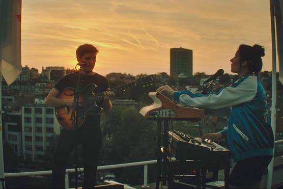 Two musicians stand on a rooftop, one holding a guitar and the other playing a keyboard, backlit by the sun setting in the background.