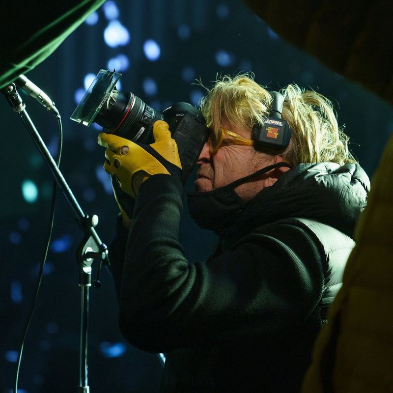 Cinematographer Anthony Dod Mantle films with a Canon camera. He is wearing a warm black jacket, yellow gloves and headphones. 