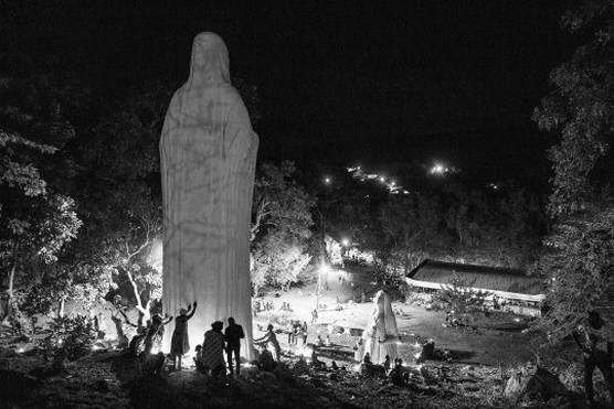 A black and white image of two enormous religious statues on a hillside with people gathered around and touching them.