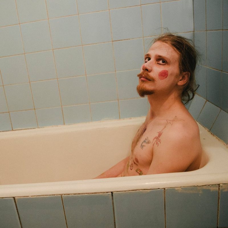 A topless man sitting in a bath wearing clown make-up.