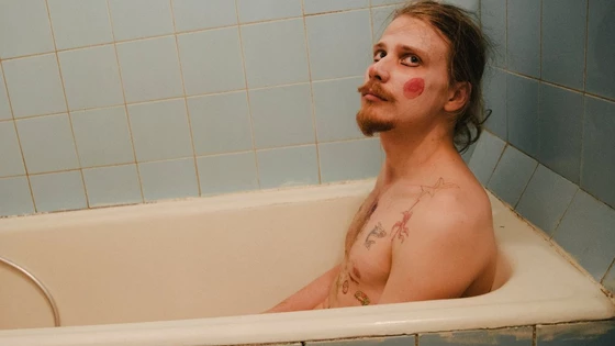 A topless man sitting in a bath wearing clown make-up.