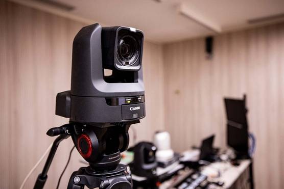 Une caméra Canon PTZ CR-N300 est fixée sur un trépied. On aperçoit un bureau à l'arrière-plan, sur lequel sont posés d'autres équipements audiovisuels.