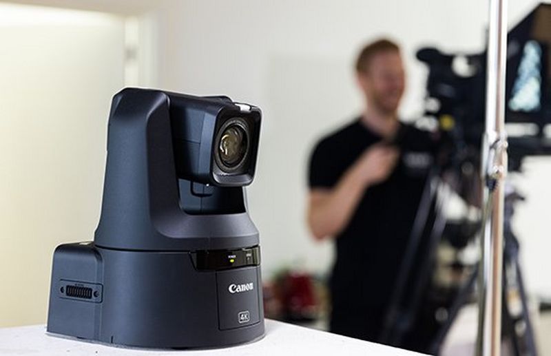 A Canon CR-N700 PTZ camera sits on a table while a man in the background operates a Canon XF605. 
