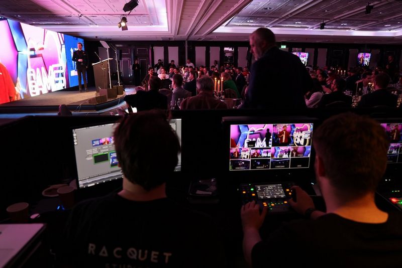 A view from behind of technicians managing multiple monitors displaying various camera angles of an awards ceremony taking place in the background.