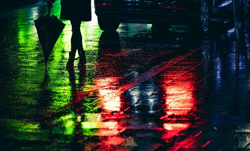Нижняя часть силуэта женщины против неоново-зеленого отражения на поверхности мокрой дороги. Красные блики стоп-сигналов автомобиля на дороге возле ее ног.
