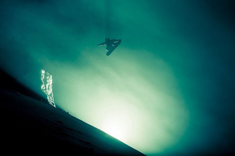 A snowboarder high in the air in foggy conditions, eerily lit from behind the crest of a hill.