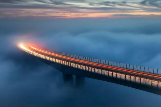 Car light trails in evening light across a curved bridge, its supports and the ground below completely obscured by thick, cloud-like fog.