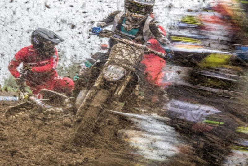 A motocross rider is caught mid race, with mud flying up into the air and other bikes blurred in action either side.