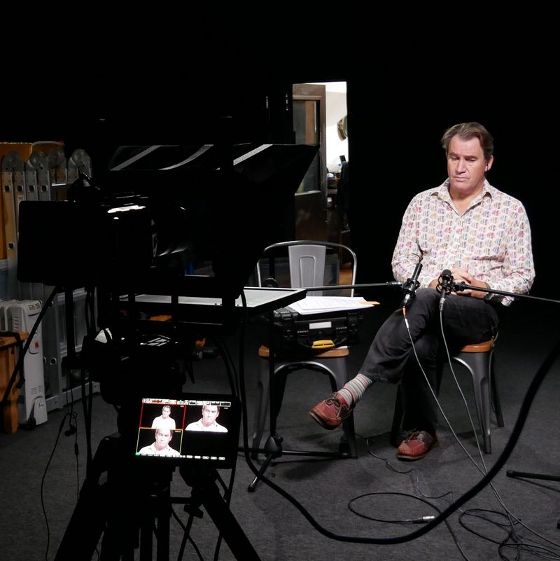 A man sits alone in a darkened studio with multiple views of him visible on the monitor attached to the camera in front of him. 