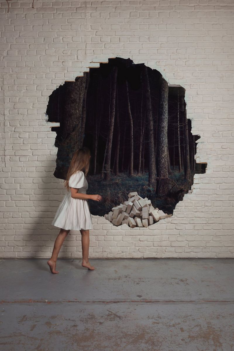 La fotografa Rosie Hardy osserva un bosco incantato attraverso un buco in un muro di mattoni dipinto di bianco in un autoritratto modificato con Canon Digital Photo Professional.