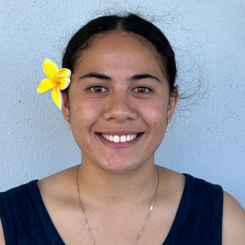 A headshot of Manaui Precious Naumalila Ash-Faulalo smiling at the camera with a yellow flower tucked behind her right ear. 