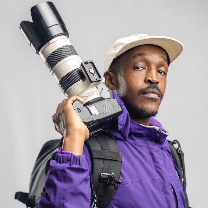 A headshot of Siyamazi Khathola wearing a purple jacket and white cap, a backpack on his back, and holding a Canon camera over his shoulder. 