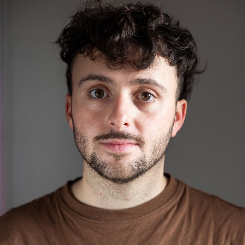 A headshot of Tomás Francisco Cuesta wearing a brown t-shirt, photographed against a grey background.