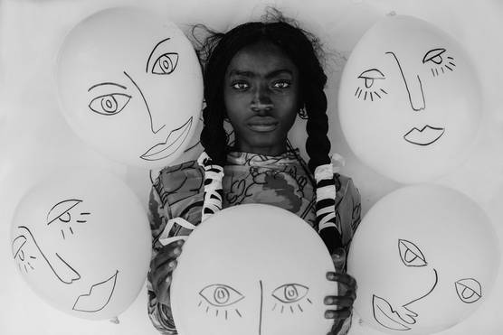A black and white portrait of a woman with braided hair surrounded by five balloons with faces drawn on them.