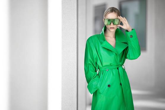 A woman wearing a green coat with her right hand in her pocket holds her left hand up to her green sunglasses while standing against a white architectural background. Taken by Sascha Hüttenhain with a Canon RF 135mm F1.8L IS USM lens.