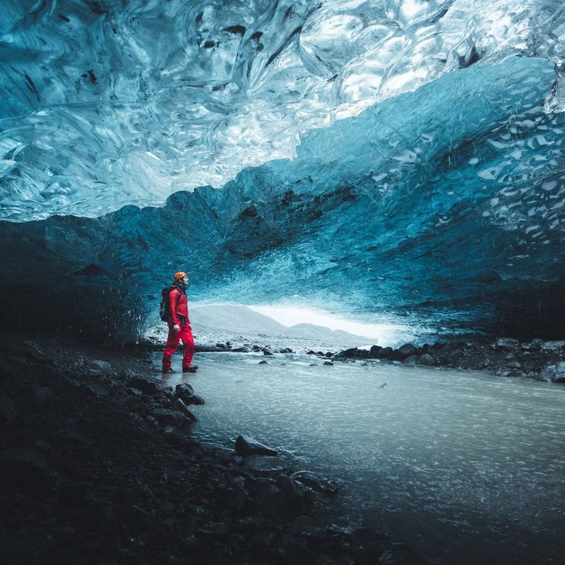 A figure dressed in red with an orange helmet stands inside an ice cave, which is varying shades of icy blue.