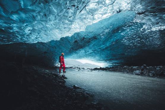 A figure dressed in red with an orange helmet stands inside an ice cave, which is varying shades of icy blue.